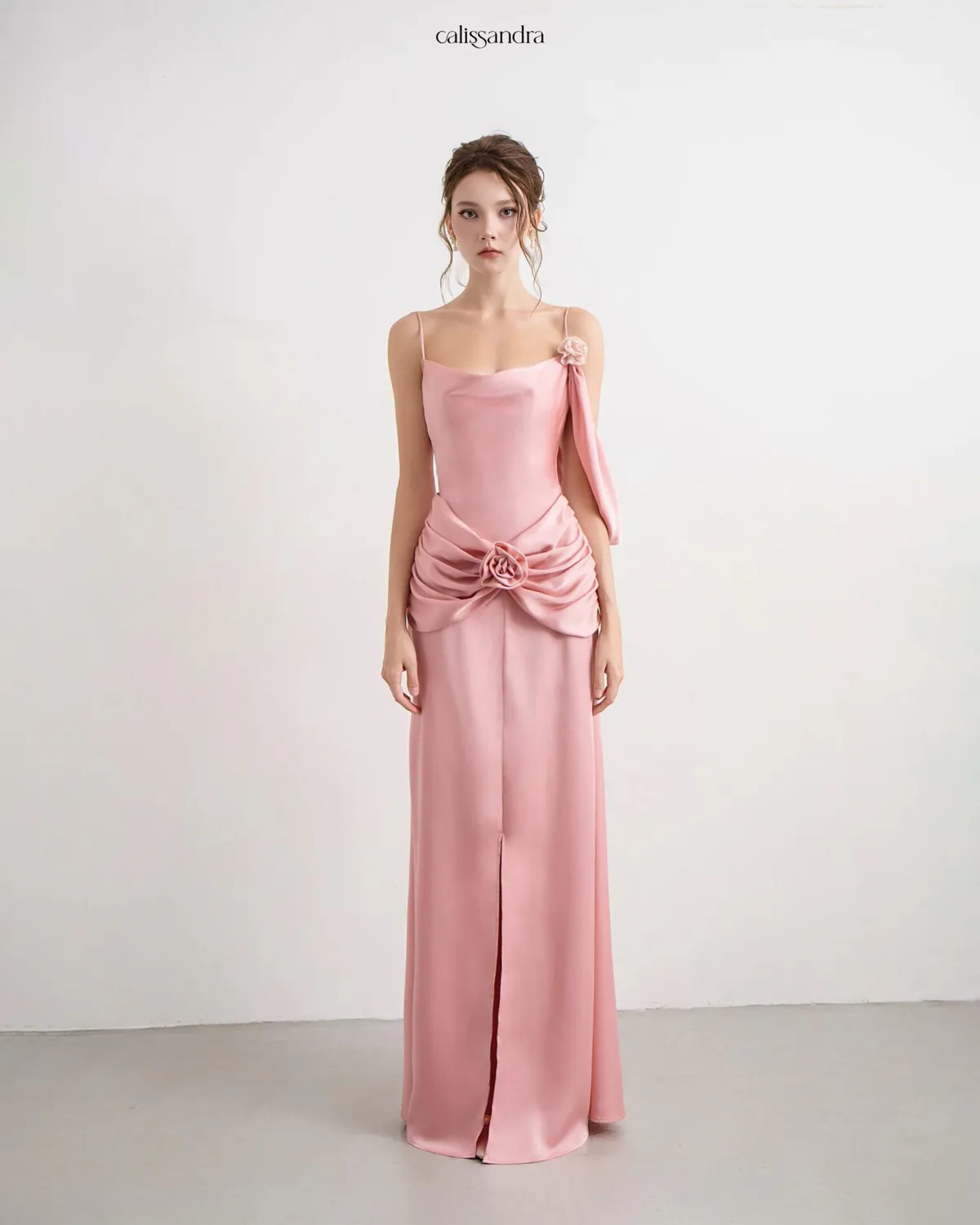 Full-length view of Auriane luxury blush pink silk satin formal evening gown, featuring a cowl neck, a front slit, and a draped waist detailed with a central 3D rose by Calissandra