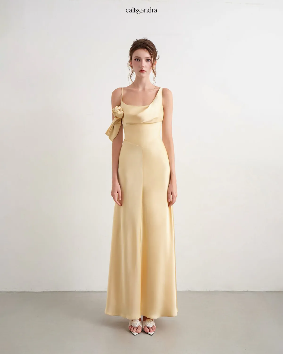 Full-length view of Roumaissa luxury butter yellow silk satin formal evening gown, featuring an asymmetrical cowl neck and a dramatic 3D rose bow accent on the strap by Calissandra