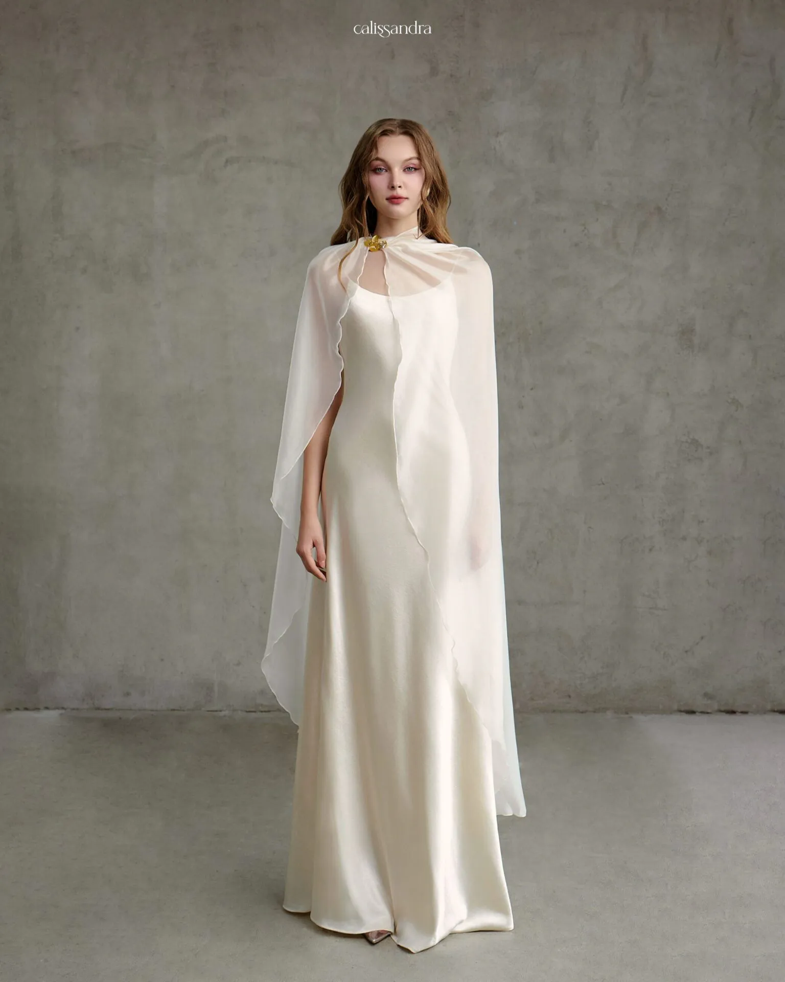 Full-length view of Ophelya luxury ivory white silk satin bridal gown, featuring a flowing sheer chiffon cape fastened with a gold floral brooch clasp by Calissandra