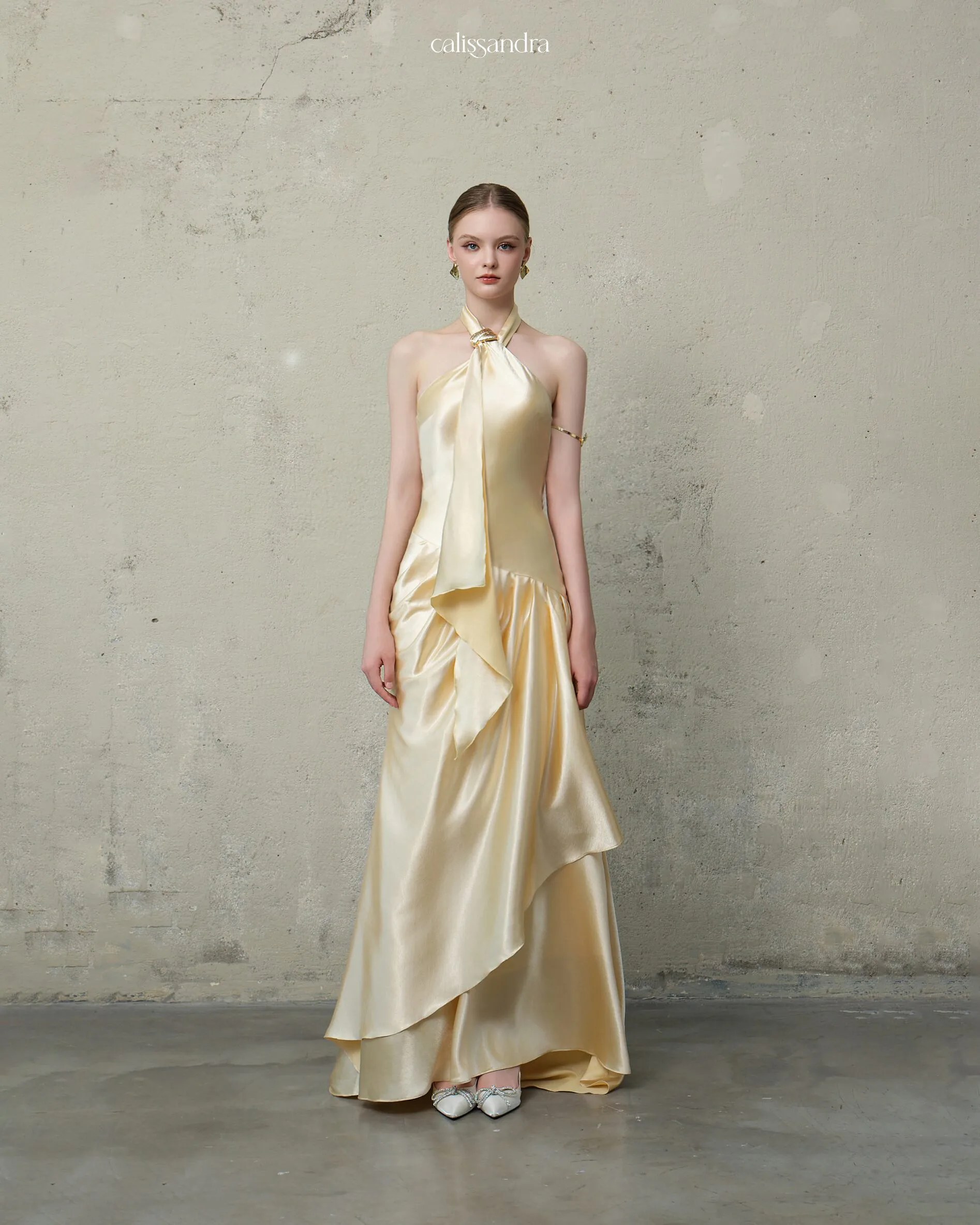 Full-length view of Ambrelle luxury champagne gold silk satin evening gown, featuring a halter neck with gold ring hardware and elegant cascading front drapes by Calissandra