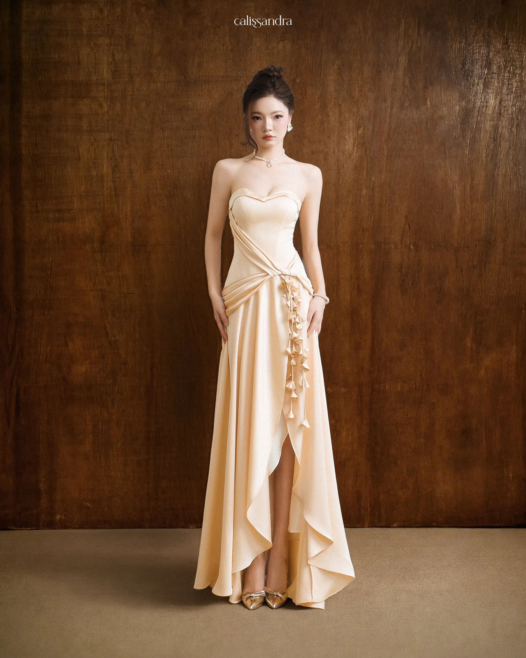 Full-length view of Estelle luxury champagne gold silk satin strapless evening gown, featuring a sweetheart neckline, a sophisticated front slit, and a cascading floral vine detail by Calissandra